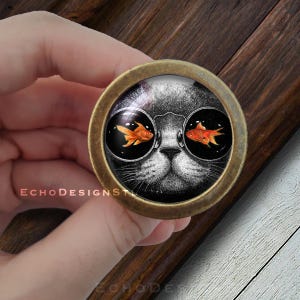 May include: A round, brass-colored knob with a black and white image of a cat wearing sunglasses. The cat's eyes are replaced with goldfish swimming in a bowl.