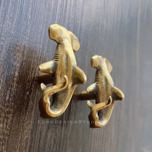 Puede incluir: Dos tiradores de armario con forma de tiburón de latón montados sobre una superficie de madera oscura. Los tiradores están detallados con aletas y cola, y son de diferentes tamaños. El texto "ECHODESIGNSTUDIO" es visible.