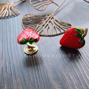 Könnte beinhalten: Zwei erdbeerförmige Dekorationsgegenstände. Einer steht aufrecht mit goldenem Sockel und Stiel, der andere liegt auf der Seite. Beide sind rot mit grünen Blättern und goldenen Akzenten. Der Hintergrund umfasst goldfarbene blattförmige Dekorationen.