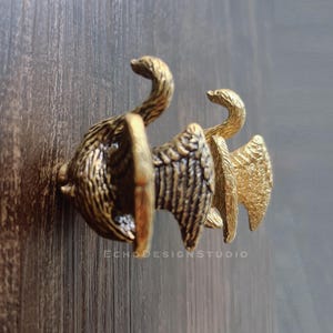 Puede incluir: Un par de tiradores de armario antiguos con forma de cisne de latón. Los tiradores presentan diseños detallados de alas y plumas, con cuellos curvos y un acabado vintage. Los tiradores están montados sobre una superficie de madera oscura.