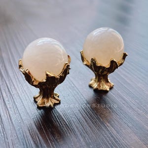 May include: Two gold decorative knobs with a round, translucent, light pink orb on top of each. The knobs have a detailed, ornate design.