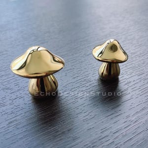 May include: Two gold mushroom-shaped cabinet knobs. The larger knob is in the background and the smaller knob is in the foreground.
