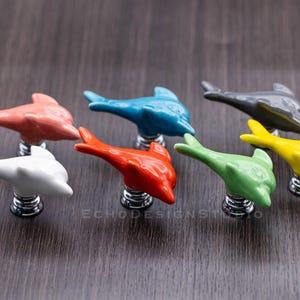 May include: Set of eight ceramic dolphin drawer pulls in various colors, including white, light blue, orange, pink, yellow, green, and gray. Each pull features a spring-loaded mechanism for easy installation.