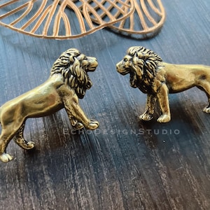 May include: Two antique brass lion figurines in a standing pose, facing each other. The lions have detailed manes and are on a dark wood surface. The text "ECHODESIGNSTUDIO" is visible.