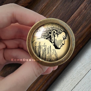 Peut inclure: Un bouton en laiton avec un dessus rond en verre représentant un dessin au trait noir et blanc d'un Bouddha. Le Bouddha est représenté de profil, la tête inclinée vers le bas et les yeux fermés.