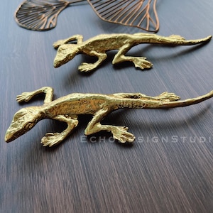 May include: Two gold-colored metal lizard figurines. The lizards are posed in a crawling position with their tails curled up. The lizards are on a dark brown wooden surface.
