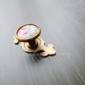 Może przedstawiać: Mosiężny gałka do szafki z kwiatowym wzorem z przodu. Gałka jest okrągła i ma ozdobną podstawę.