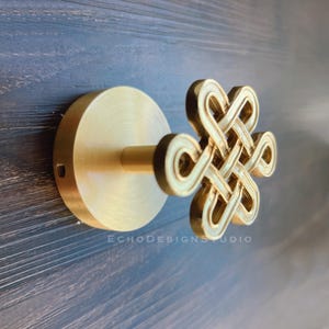 Może przedstawiać: Złoty metalowy haczyk ścienny z ozdobnym wzorem węzła. Hak jest zamontowany na okrągłej podstawie.