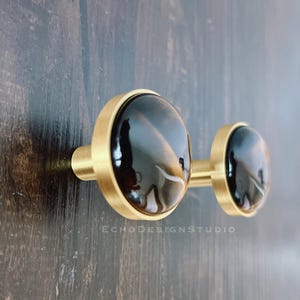 Peut inclure: Deux boutons de cabinet dorés avec des incrustations d'agate noire. Les boutons sont ronds et ont une finition lisse et polie.