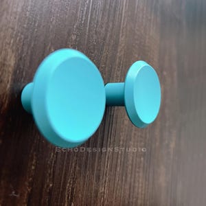 Puede incluir: Dos pomos redondos de color turquesa azul para armario con un borde ligeramente elevado. Los pomos están montados en una superficie de madera oscura.