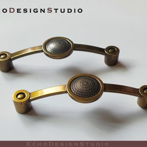 May include: Two antique brass cabinet pulls with ornate circular designs. The pulls have a slightly curved shape and are designed for use on drawers or cabinets.