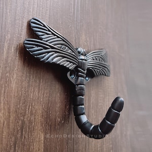 May include: A dark grey metal dragonfly hook fixed to a wooden surface. The hook is shaped like a dragonfly with detailed wings and a curved body. The text "ECHODESIGNSTUDIO" is visible on the hook.