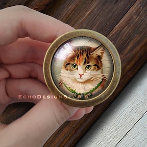May include: A vintage-style, round, brass-colored knob with a glass insert featuring a portrait of a tabby cat wearing a green and red holly garland with bells.