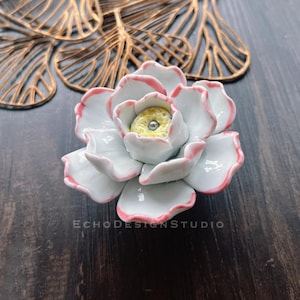 May include: White ceramic flower-shaped cabinet knob with a pink rim and a yellow center. The knob is on a dark wood surface.