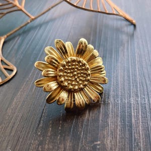 Może przedstawiać: Złoty metalowy pierścionek w kształcie słonecznika z fakturowanym środkiem. Pierścionek leży na ciemnym drewnianym tle.