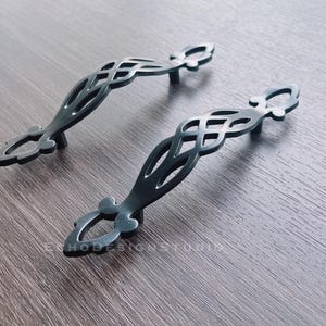 May include: Two black metal cabinet door pulls with an intricate, woven design. The pulls have a decorative, ornate finish.