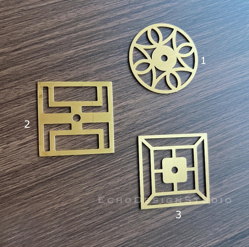 May include: Three brass metal filigree shapes: a circular design with a central hole, a square with an open geometric pattern, and a square with a central square and a geometric border. The shapes are on a dark wood surface.