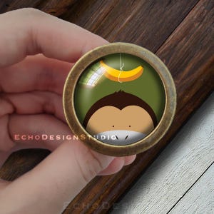 Op de afbeelding: Een ronde, messingkleurige knop met een glazen inzetstuk met een cartoon-aap met een banaan die boven zijn hoofd hangt. De aap is bruin met een groene achtergrond.