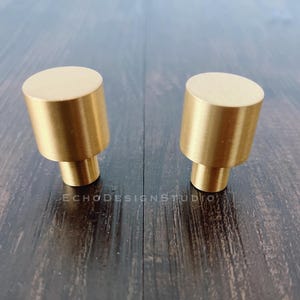 May include: Two gold-toned metal cabinet knobs with a cylindrical shape. The knobs have a smooth, polished finish.