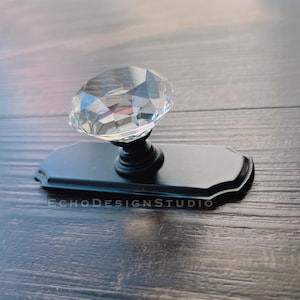 Pode incluir: Uma base retangular preta com um botão de cristal transparente em forma de diamante. O botão é facetado e reflete a luz. A base tem uma borda decorativa e o texto "ECHODESIGNSTUDIO" está impresso na base.