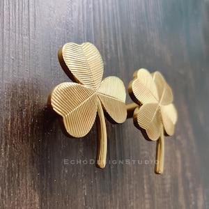 May include: Two gold-toned metal clover leaf shaped cabinet knobs. The knobs are attached to a wooden surface.