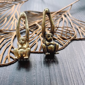 Puede incluir: Dos figuritas de mono doradas con los brazos levantados en el aire. Los monos están de pie sobre una superficie de madera con un diseño de hoja de oro.