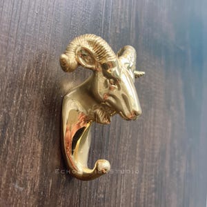 Op de afbeelding: Een messing haak in de vorm van een ramshoofd, gemonteerd op een houten oppervlak. De haak is goudkleurig en heeft een gedetailleerd ramshoofd met gebogen hoorns en een gebogen haak om items op te hangen. Een decoratief en functioneel item.