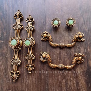 Può includere: Set di cinque pomelli per armadi in stile antico con lavorazione in metallo decorata e inserti in vetro verde. I pomelli sono di varie forme, tra cui due tiretti lunghi, due tiretti più piccoli e un pomello rotondo.