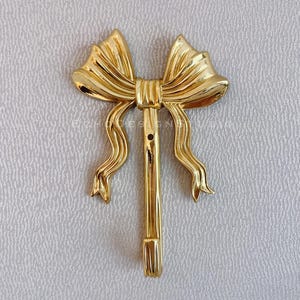 Może przedstawiać: Złoty metalowy haczyk w kształcie kokardki z ozdobnym wzorem. Haczyk jest przymocowany do ściany.