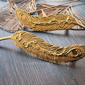 May include: Two gold-coloured feather-shaped decorative objects with detailed patterns. The feathers have a textured surface and floral accents. A matching gold-coloured, openwork leaf design is in the background.