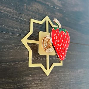 Può includere: Un pomello decorativo per mobili raffigurante una fragola rosso brillante con foglie verdi e semi bianchi. La fragola è fissata su una base circolare dorata montata su una piastra posteriore geometrica a forma di stella color oro con il testo ECHODESIGNSTUDIO.
