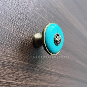 Puede incluir: Tirador de armario turquesa y latón. El pomo redondo presenta una cara turquesa, un borde de latón y un vástago de latón. Montado sobre una superficie de madera oscura, ideal para muebles.