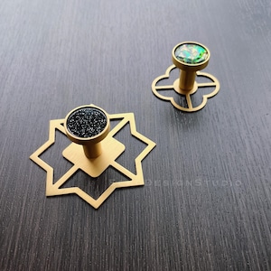 Puede incluir: Dos pomos de armario dorados con dise&ntilde;os geom&eacute;tricos. Un pomo tiene una parte superior de purpurina negra y el otro tiene una parte superior de purpurina verde.