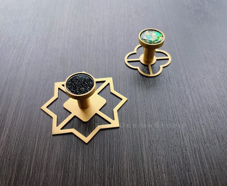 Puede incluir: Dos pomos de armario dorados con dise&ntilde;os geom&eacute;tricos. Un pomo tiene una parte superior de purpurina negra y el otro tiene una parte superior de purpurina verde.