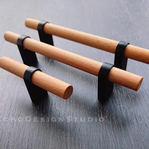 Op de afbeelding: Drie houten kastgrepen met zwarte metalen accenten. De grepen zijn van verschillende grootte en zijn in een rij op een donker houten oppervlak gerangschikt.