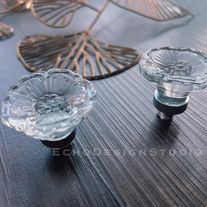 Könnte beinhalten: Zwei klare, blumenförmige Schrankknöpfe aus Glas mit silbernen Metallsockeln. Die Knöpfe befinden sich auf einer dunklen Holzoberfläche.