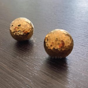 Puede incluir: Dos bolas de metal martillado de color dorado. Las bolas tienen una superficie rugosa y texturizada.