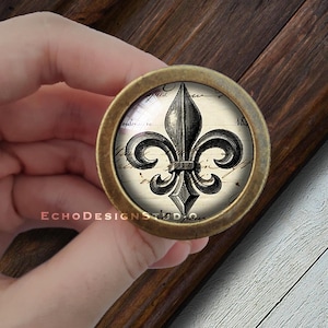 Peut inclure: Un bouton rond de couleur laiton avec un insert en verre représentant une illustration vintage en noir et blanc d'une fleur de lys sur un fond de texte manuscrit.