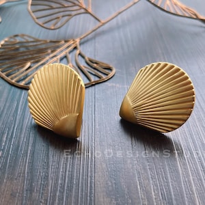 May include: Two gold-coloured, seashell-shaped cabinet knobs with a fan-like design. The knobs are on a dark blue wooden surface, with a decorative gold leaf in the background. These knobs are likely for furniture.