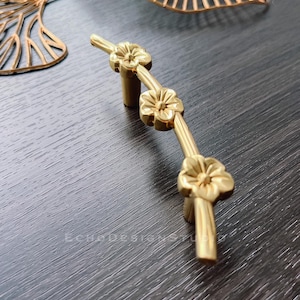 Könnte beinhalten: Goldfarbene Schrankbeschläge mit floralem Design. Die Beschläge haben drei stilisierte Blüten auf einem gebogenen, astförmigen Stiel.