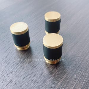 Può includere: Tre pomelli per armadi di colore dorato con accenti neri. I pomelli sono rotondi e hanno una finitura liscia e lucida.