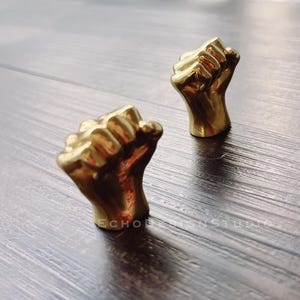 以下が含まれることがあります： 金色の金属製の拳の彫刻が2つ。拳は空中に突き上げられており、親指は鑑賞者の方向を向いています。