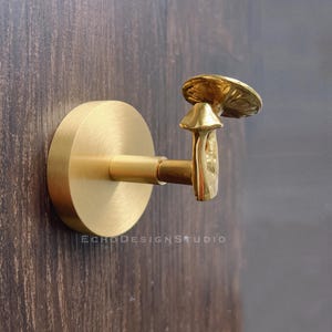 Op de afbeelding: Gouden wandhaak in de vorm van een paddenstoel met een ronde basis. De haak is bevestigd aan een houten muur.