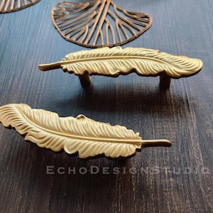 May include: Two gold-toned feather-shaped cabinet pulls. The pulls are made of metal and have a detailed design.