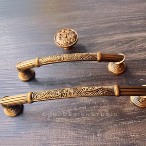 May include: Ornate bronze-toned cabinet hardware set. Includes two long pull handles and a round knob, all with detailed floral and leaf patterns. The hardware is set against a dark wood grain background. The handles are approximately 20 cm long.