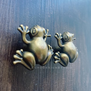 May include: Two bronze-colored frog-shaped cabinet knobs. The knobs are detailed with textured skin and expressive eyes, giving them a whimsical appearance. The knobs are mounted on a dark wood surface.