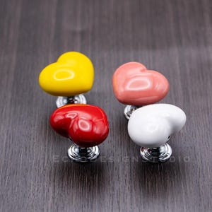 Puede incluir: Cuatro pomos de armario de cerámica en forma de corazón en amarillo, rosa, rojo y blanco. Los pomos tienen un acabado plateado.