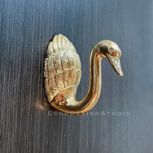Puede incluir: Un gancho dorado en forma de cisne con plumas detalladas en la parte posterior. El gancho está montado en una superficie de madera oscura.