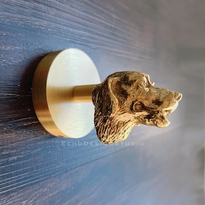 Può includere: Un gancio da parete in metallo dorato a forma di testa di cane. Il gancio è montato su una base rotonda e ha una finitura testurizzata.