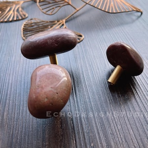 Puede incluir: Dos pomos de armario de piedra marrón con detalles en latón. Los pomos tienen forma de piedras redondeadas y tienen una varilla de latón que conecta las dos piedras.